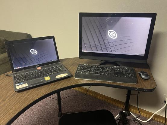 Two computers on a curved desk. One a laptop, another an "all in one"  computer built with screen. The desktop shows the Linux Mint distribution logo.