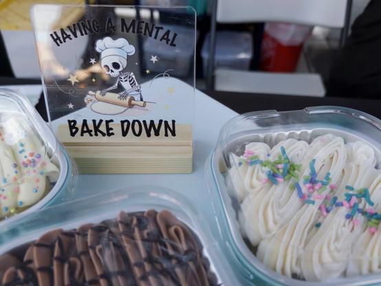 A humorous sign reading “HAVING A MENTAL BAKE DOWN” sits prominently in the center of the image, featuring a cartoon skeleton wearing a chef’s hat while rolling dough on a cloud surrounded by stars. The sign is positioned between glass containers of frosted cupcakes - chocolate cupcakes with dark frosting on the left and vanilla cupcakes with white buttercream frosting and colorful sprinkles on the right.