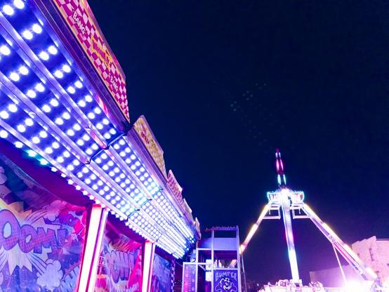 Brightly lit carnival rides dominate a nighttime scene against a dark sky. In the foreground, an ornate ride with intricate LED light patterns in white and purple creates geometric designs along its decorative facade. To the right, a large pyramid-shaped ride structure rises into the darkness, outlined in white lights with red accents at its peak. The dramatic upward angle of the photograph emphasizes the towering scale of these illuminated attractions, creating a vibrant contrast between the artificial carnival lighting and the dark evening sky. The scene captures the electric energy and nostalgic atmosphere of a traditional fairground at night.​​​​​​​​​​​​​​​​