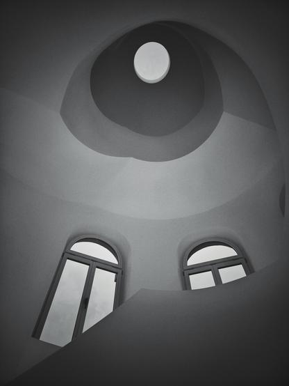 A portrait-orientated monochrome, low-angle shot looking upwards within a structure, possibly a dome or a rotunda. The ceiling features a circular oculus at its apex, through which a light source, likely the sky, is visible. Below the oculus, the ceiling recedes downwards in concentric, circular steps, creating a spiraling or tiered effect.
Further down and closer to the viewer, two arched windows are visible. The window on the left appears slightly larger and positioned lower than the one on the right. Both windows reflect what appears to be an overcast sky. The overall tone of the image is dark, with a vignette effect that darkens the corners and edges of the frame. The surfaces of the structure appear smooth and unadorned.