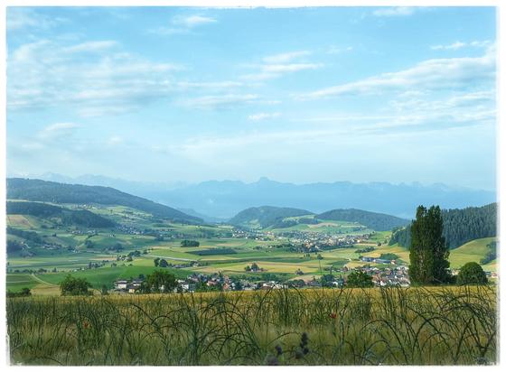 A wide valley with fields and villages. In the back some hills with forest and in the mist a range of mountains.