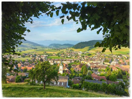 Picture framed by branches of some trees. A village a the foot of a hill, behind it hills and mountains at the horizon.