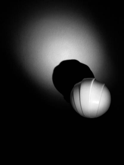 Black and white  photo of a round white glass top of an old lamp. The picture was taken in the dark with the lamp off, hwile the phone camera had its flash on, casting a distinct black shadow on a wall. As well, surrounding the shadow is a diffuse oval shaped area lit up by the flash.