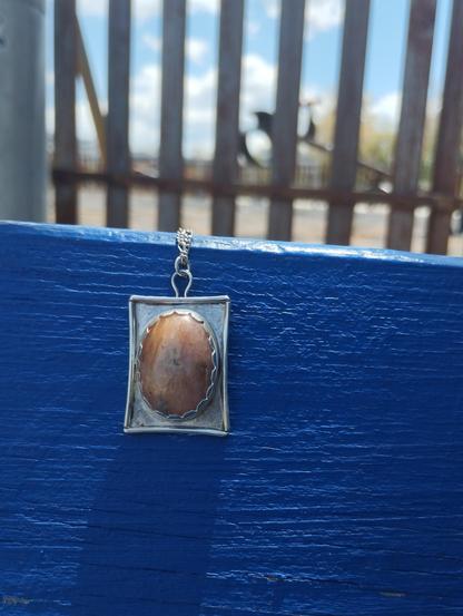 a pendant, it has an oval stone in silver rectangular frame. It's placed on a blue painted wood