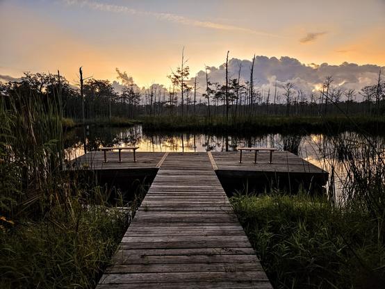Pre-Sunrise glow over a wetland landscape and waterway beyond an old wooden observation deck with bench seating. Enormous cloud formations hover at the horizon in dark grey shades creating the illusion of a mountain range beyond the field of grasses, reeds, and barren trees. Orange and yellow glowing shades rise from behind the clouds as sunrise approaches, illuminating the waterway and the marsh land.