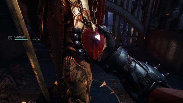 **TL;DR:** Close-up from *Doom: The Dark Ages* showing the player grabbing a large red gemstone from an armored figure, with “PLUNDERER” progress displayed.

This image is a dramatic first-person close-up from *Doom: The Dark Ages*. The player, wearing a spiked gauntlet, is grasping a large, blood-red, heart-shaped crystal set in a golden clawed frame, hanging on a thick chain around a heavily armored humanoid figure. The background shows gothic architecture with barred gates. On the left, the HUD displays a task labeled “PLUNDERER” with a progress bar reading 124/150. The scene emphasizes looting or acquiring a key item in the game.