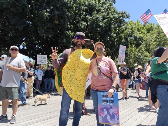 A man wearing a bright yellow taco costume, smiling and holding up a peace sign, next to a person smiling holding a protest sign and wearing a shirt with a pastel rainbow flame and fist that says "Stonewall Rebellion '69". Behind them is a crowd of protesters with signs.