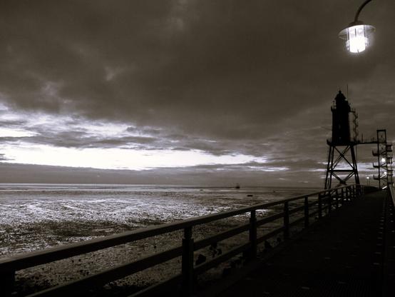 Black and white photo. It is evening by the sea. On the right of the picture, the light of a lamp shines from above onto a wooden walkway leading to the Obereversand lighthouse. It is a black, four-sided tower on a lattice construction, with two galleries and a round lantern. The mudflats stretch out to its left. The sky is mostly cloudy.
