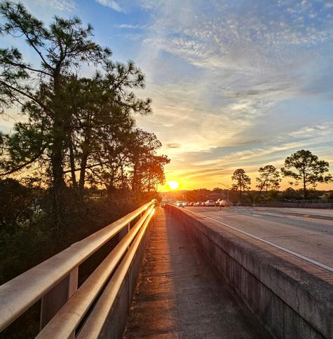 Morning sunrise viewed climbing a sloped pedestrian bridge, with the big golden sun sending a beam of light down the pathway ahead, reflecting off the metal hand rails, and filling the horizon with warm morning shades of yellow and orange beneath the big blue Florida sky.