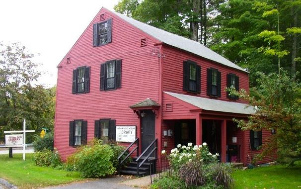 An old house painted red with black shutters, surrounded by trees. There is a sign next to the door and on the lawn: "Salmon Falls Library". There are flowers in front of the building.