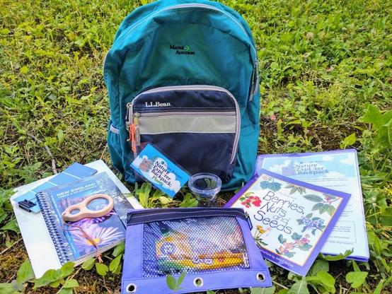 A teal backpack with LL Bean / Maine Audobon logo. The contents of the backpack are spread on the ground, and includes guidebooks, a magnifying glass, and other items for exploring the outdoors.