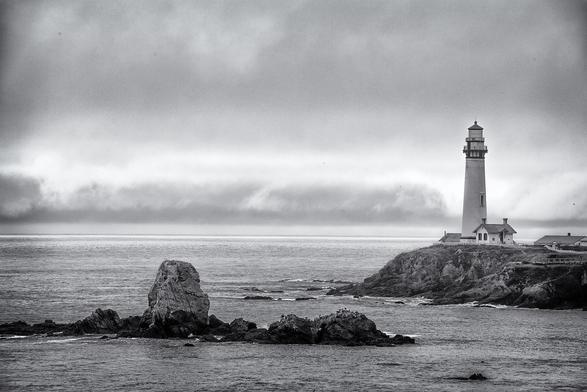 Black and white photo of Pigeon Point lighthouse on a foggy morning. In addition to the lighthouse, we see a rocky outcropping offshore.