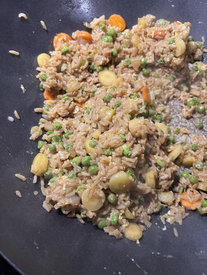 Photo of rice in the bottom of a wok, getting fried up. Mixed in are bits of carrot, onion, peas, and garlic scapes.