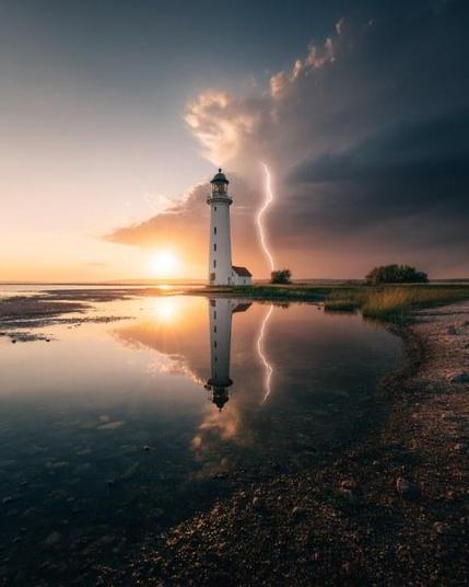 Lightning strikes parallel to a lighthouse, with the sun at the horizon. Both are reflected in the water.