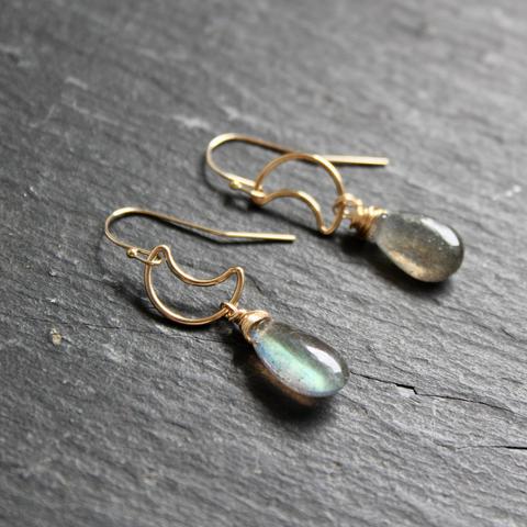 A photo of a pair of handmade blue and gold dangle earrings on a dark grey slate background. Earrings are made of 14ct gold-filled wire, 14ct gold-filled crescent moons, and teardrop-shaped labradorite stone beads.