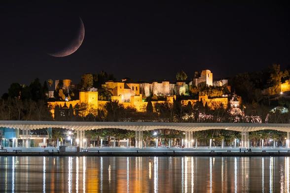 Una vista de El Paseo de las Palmeras y la Alcazaba de Málaga de noche con una enorme luna.