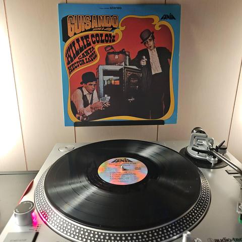 A vinyl record sits on a turntable. Behind the turntable, a vinyl album outer sleeve is displayed. The front cover shows Willie Colon and Hector LaVoe near an open safe. One is leaning against the safe holding a gun, the other is sitting near it counting money