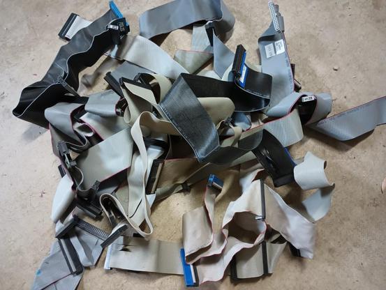 A large, around a foot wide, tangled pile of old computer ribbon cables in gray and black colors. Some of the end connectors are blue but most are black. The pile lies on a concrete floor.