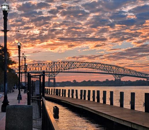 Rosy sunrise across a very long, wooden fishing pier with an enormous, arched steel bridge in the distance,  all illuminated with soft pinkish red light beneath blue sky overtaken by countless overlapping fluffy clouds of blue, white, and grey.