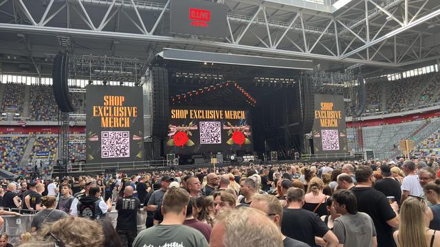 The image depicts a large outdoor concert in a stadium setting. In the foreground, a dense crowd of people is gathered, many wearing casual concert attire, with some individuals wearing black shirts. The crowd is facing a large stage at the center of the image, which is equipped with multiple large screens displaying the text "SHOP EXCLUSIVE MERCH" in bold yellow letters, accompanied by QR codes. The screens also feature a graphic of a rose with thorns. The stage is set up with professional lighting and sound equipment, and the structure of the stadium is visible, with a metal framework and a partially covered roof. The stands are partially filled with spectators, and the overall atmosphere suggests a lively music event.

Provided by @altbot, generated privately and locally using Ovis2-8B

🌱 Energy used: 0.177 Wh

Übersetzen