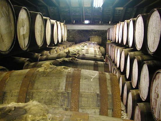 A bonded warehouse at Glenmorangie Distillery. The image shows a view across the tops of a line of whisky casks. There are more rows of casks, stacked three high, on the left and right of the image. Some light is coming in through a roof light at the far end, above a stone wall.