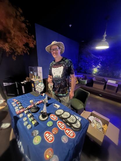 Me in a tie dye shirt standing behind a blue table with stickers. Purple hue background.