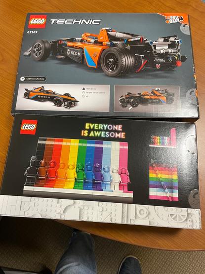 Two unopened LEGO sets on a desk.  The top set is a model of a McClaren Neom Formula e racing car.  The bottom set is the Everyone Is Awesome set for Pride.