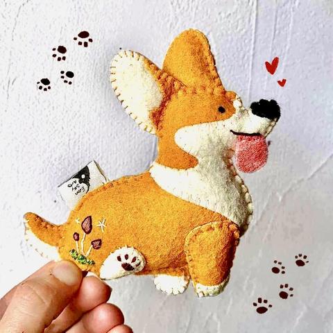 Picture of a handmade felt plushie held in front of a white wall. It is standing to the side, it’s ears are big and orange like it’s body, their inside are ivory, like it’s face, neck and feet. 
The corgi’s hind legs are facing up and the bottom of its feet are visible. It has a small tail and a big tongue coming out of its embroidered mouth. Three mushrooms are embroidered on its butt.