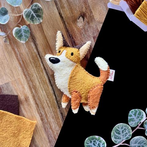 Picture of my most recent corgi plushie photographed in front of a black and wooden background, surrounded with decorative plants and matching felt and threads. Its ears are a dark brown with a cream-coloured inside. Its body is a soft orage and its muzzle and belly a soft ivory. Two small beady eyes are embroidered on the same side as well as one big, shiny black nose. Its legs and tail are a darker soft orange with ivory tips.