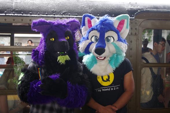 Two people with fursuit heads posing in front of the side of a BMT Standard train. On the left, the person (Instagram @J.J2644) is wearing a fursuit head and fursuit claws with purple & black fur and a "ty" tag hanging on the left ear. On the right, the person (Instagram @ramseybrony17) is wearing a cartoonish fursuit head with more light-coloured fur and big eyes, as well as a shirt with the yellow service bullet (the circular icon) for the Q train with the words "Coney Island to 2 Av".