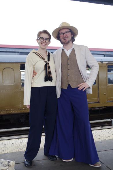 Two people in rather casual outfits posing in front of the side of a BMT Standard train across the tracks. On the left, the person (@esteoli.bsky.social) is wearing a white blouse and dark blue trousers, with a brown-white-black sailor tie. On the right, the person (@aaronblanton.bsky.social), wearing a light tan jacket, a darker tan cardigan, a light blue trouser and a cowboy hat (?), is holding the person on the left by the waist.