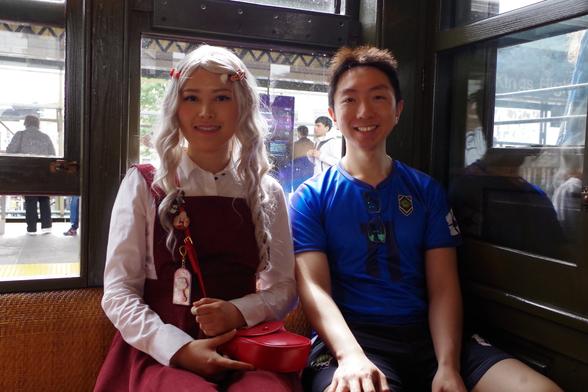 Two people going to an anime convention (who mistakenly boarded this train...) seated on a BU car. On the left is someone cosplaying as Eri in My Hero Academia, with a long white wig, a white blouse, a red jumper skirt and a red purse, with character memorabilia on the belt of that purse. On the right is a person in a blue soccer shirt. Both smiling.
