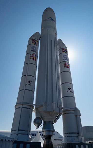 A full-size model of the giant Ariane 5 rocket, with its core stage and twin boosters. The rocket is white with flags and logos from ESA, CNES, and the member states in the Ariane programme. The sun is behind the rocket.