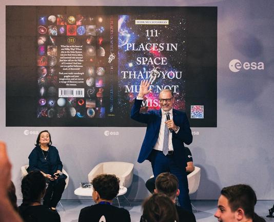 A picture of a man in a blue suit standing with one hand in the air in front of an LED screen showing a book covering. There are kids in the audience in the foreground.