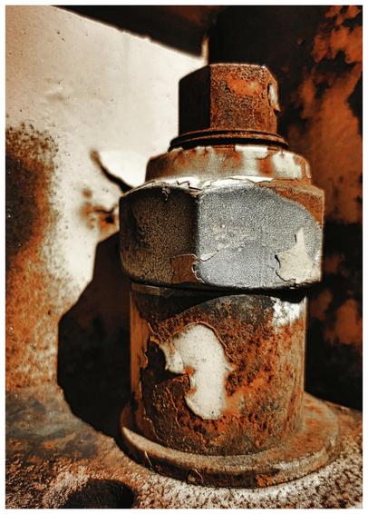 Das Bild zeigt einen stark verrosteten, zylindrischen Bolzen aus Metall im Detail. Die Rostschicht ist dick und uneben, mit großen, blätternden Stellen, die die darunterliegende metallische Oberfläche zum Vorschein bringen. Die Farbe des Rosts variiert zwischen tiefem Orangebraun und einem helleren, fast weißen Farbton, was auf unterschiedliche Rostoberflächen und -stadien hinweist. An einigen Stellen ist die ursprüngliche Farbe des Metalls, ein matter Grauton, noch sichtbar. Der Bolzen ist mit zwei deutlich sichtbaren Muttern versehen, wobei die obere Mutter ebenfalls stark vom Rost befallen ist. Der Hintergrund des Bildes ist unscharf, aber man erkennt eine ebenfalls verwitterte und vom Rost befallene Oberfläche.