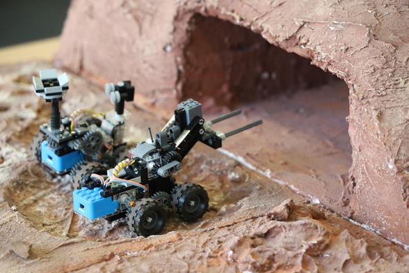 Sitting side by side on rough red model terrain, two small four-wheeled Lego rovers are facing a tunnel-shaped opening in the model landscape. It looks like they are about to start racing each other.