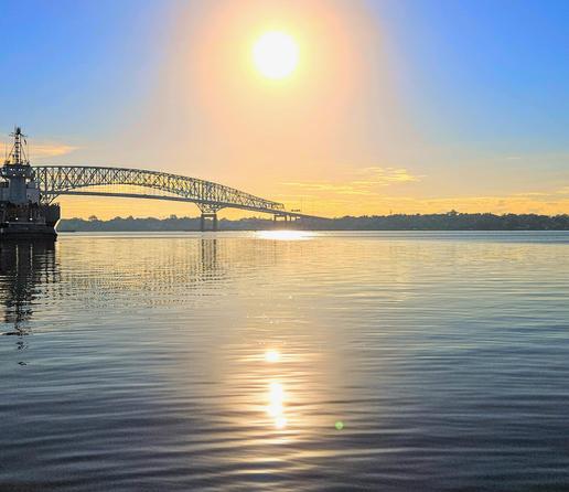 Brilliant, blazing sun rising in clear blue morning sky over a vast shimmering river with an enormous arched bridge in the background, all casting their reflections upon the water below.