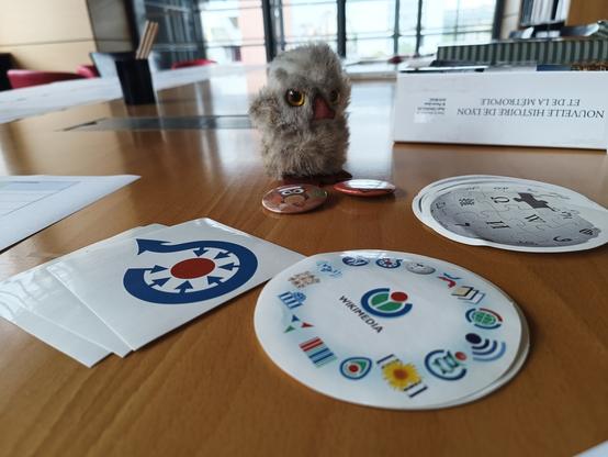 Photographie numérique couleur en format paysage.
Sur une table en bois vue de près et en perspective, des autocollants avec les logos de Commons (la médiathèque de Wikipédia), Wikimédia et Wikipédia. 
Il y a également une petite chouette en peluche et 2 badges chouette, ainsi que des ouvrages posés, dont la nouvelle histoire de Lyon et de la Métropole.
Au fond, une baie vitrée et des fauteuils rouges.