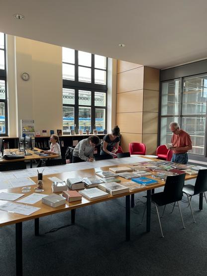 Photographie numérique couleur en format portrait.
Vue de la même table vue de plus loin, avec l'ensemble des ouvrages préparés, un homme qui tient un livre, 2 personnes penchées sur les listes des images à intégrer et une autre entrain de travailler sur un ordinateur.
A l'arrière-plan, des fenêtres donnant sur la gare avec un train bleu à quai, et une partie de la baie vitrée.