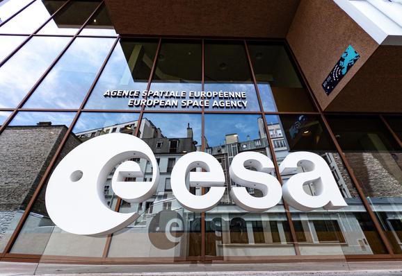 A picture taken close to ground level in front of the ESA HQ building in Paris. There is a large white 3D ESA logo in the foreground, with a glass facade behind reflecting buildings and the sky.