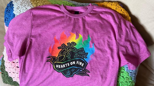 A pink-purple, berry-colored Denver Pride t-shirt draped across a folded, crocheted afghan. The shirt design has a black anatomical heart surrounded by rainbow colored fire with the words “Hearts on Fire” in the middle.