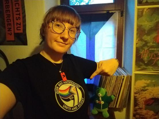 Ich, große runde Brille, Ponyfrisur, mit einem Shirt der QueerPride vom letzten Jahr, mit Flaggen und dem Spruch "Queer and Antifascists Unite and Resist"