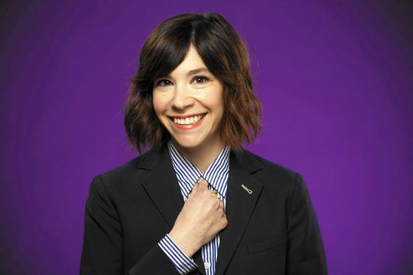 Headshot of Carrie Brownstein smiling: a beautiful brunette, Brown eyed, white woman with short wavy hair wearing a black suit jacket & blue/white striped button down shirt.