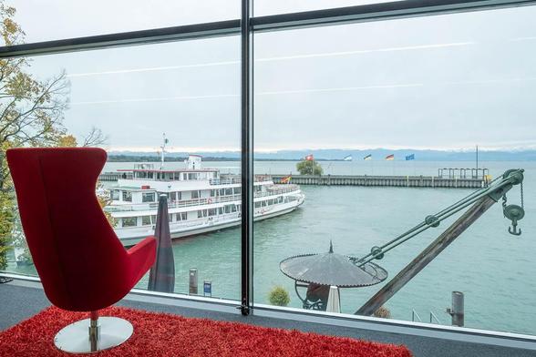 A modern red chair on a red rug sits by a large window overlooking a docked white passenger boat, with calm water and a pier in the background under an overcast sky.