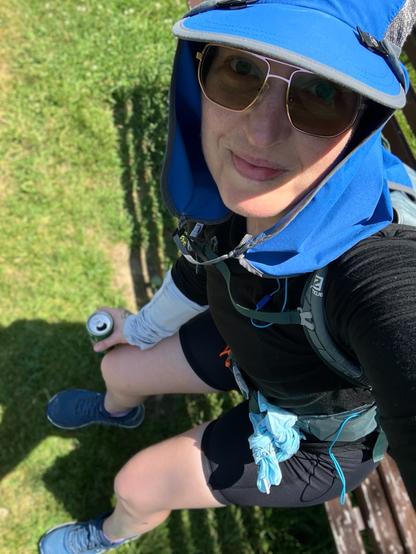 A middle aged white woman, mid run, sitting on a park bench having a soda pop. She is clad in running clothes with sunglasses and is smiling at the camera.