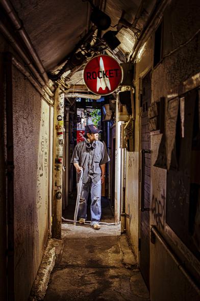 A man is crossing a dark narrow alley in Golden Kai, Shinjuku, Tokyo, Japan. His face and body are illuminated by articifial light coming from an open door on the side of the alley. The frame around him is just slightly bigger than his body.