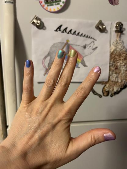 A hand held up, palm facing away so as to display the rainbow-painted nails (from pinky to thumb: blue, turquoise, lime, pink, lilac). In the background is a refrigerator, including some fridge art of a screaming opossum carrying a progress pride flag.