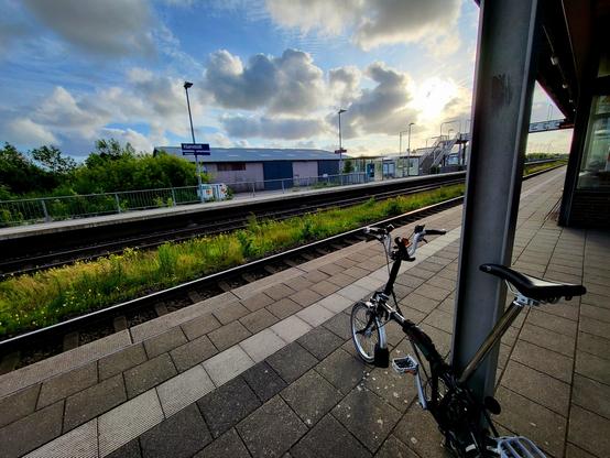 Ein Brompton Fahrrad angelehnt an einem Stahlträger auf einem Bahnsteig.  Auf der anderen Seite des Gleisbett weist ein blaues Haltestellenschild auf Klanxbüll hin. Darüber freundlicher blauer wolkenbehangener Himmel mit Abendsonne.