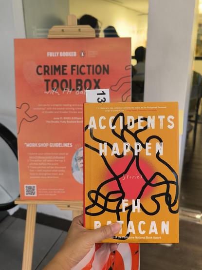 hardbound copy of accidents happen by fh batacan the cover has outlines of bodies as if in a crime scene, behind it is the poster for the author's signing session