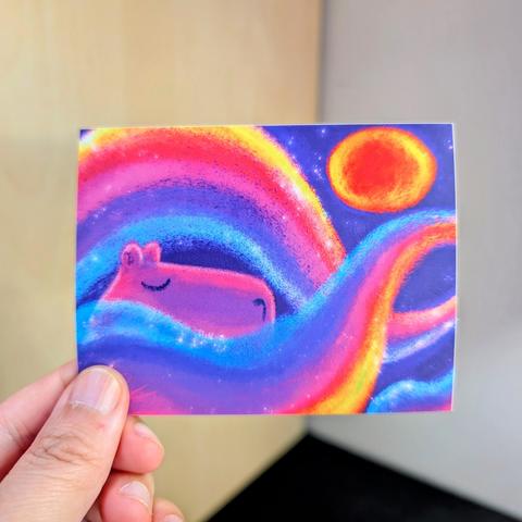Photo of a left hand holding a rectangular sticker. The sticker is a matte painting of a capybara sleeping among rainbows.